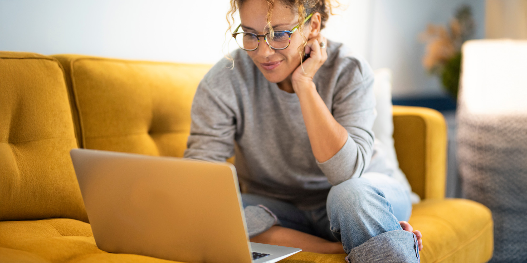 A lady sitting on a yellow couch with her laptop look at something that makes her happy.