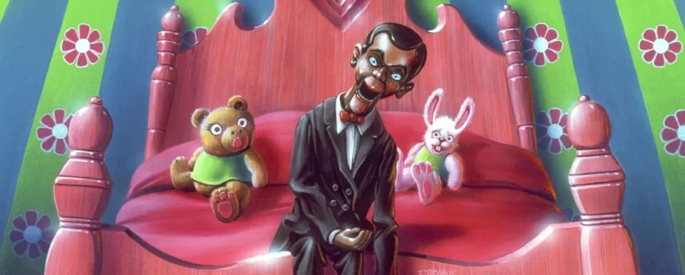 Slappy from Goosebumps sitting on a pink bed with a teddy bear and bunny plushie sitting next to him.